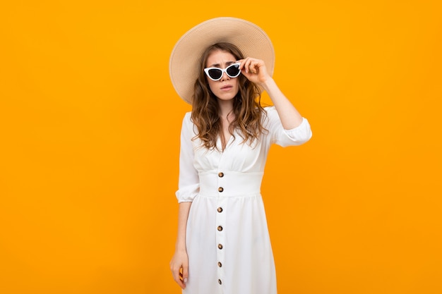 Fashionista tourist girl in a straw hat with sunglasses dressed in a white dress on a yellow background with copy space