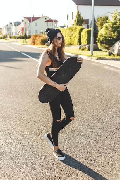 Fashionly dressed skater girl posing with board on the city road. Sunset time.