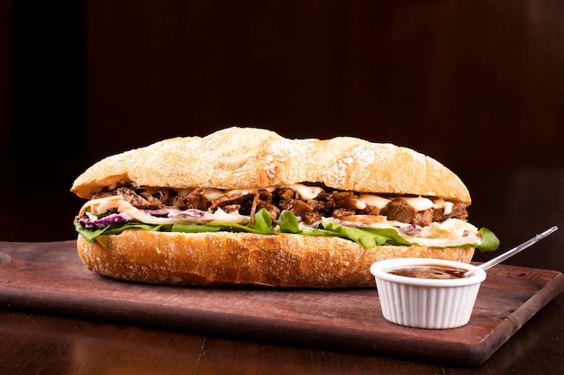 Fast food beef brisket sandwich with arugula and coleslaw salad on baguette bread on wooden board with barbecue sauce dark background from the front