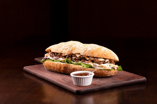 Fast food beef brisket sandwich with arugula and coleslaw salad on baguette bread on wooden board with barbecue sauce dark background