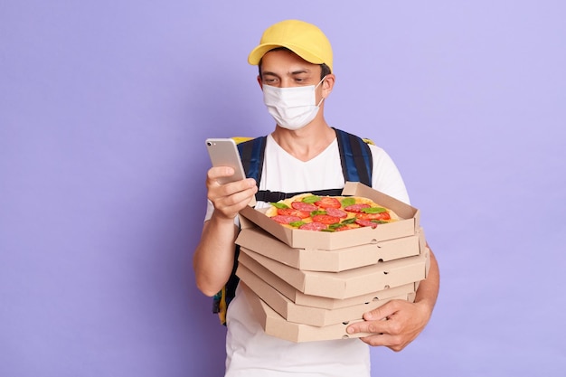 Fast service and best food delivery Kind pleased male courier wearing protective mask holding pizza boxes from restaurant and using smartphone isolated on purple background