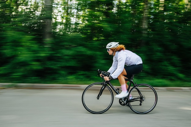 Fastmoving redhead woman with curly hair in a helmet riding street racing bike on a road Overgrown trees by the sides with motion blur