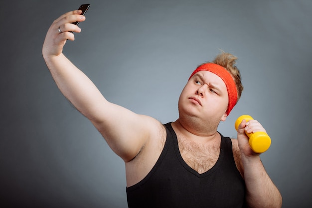 Fat man with big belly, holding dumbbell, doing selfie on grey wall