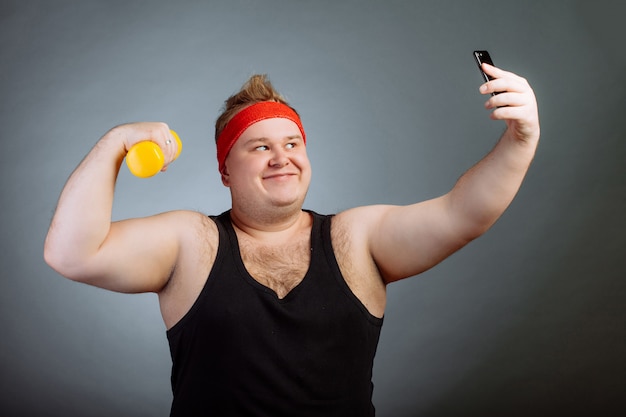 Fat man with big belly, holding dumbbell, doing selfie on grey wall