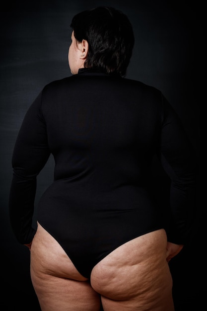 A fat woman with cellulite on her buttocks stands with her back against a black background