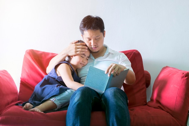 Father and daughter are reading books and she is sleepingSitting on a red sofaIt is a good learning practice and a good family relationship