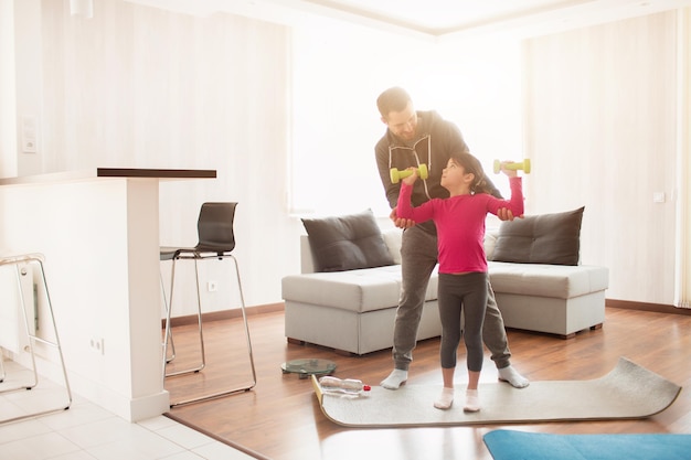 Father and daughter are training at home. Workout in the apartment. Sports at home. They are standing on a yoga mat. Girl holding dumbbell.