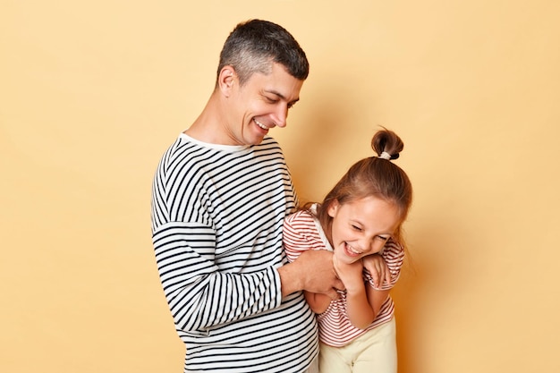 Father and daughter having fun together isolated over beige background family wearing striped shirts dad tickles little kid and laughing happily