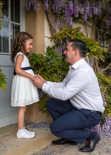 Father and daughter A little girl in a white dress is standing dad kneeled in front of her Look into each other's eyes Father's love and care for his daughter Flower background wisteria