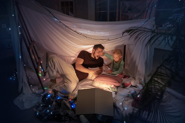 Father and daughter sitting in a teepee, having fun, playing with the flashlight in dark room with toys and pillows. Look happy. Home comfort, family, love, Christmas holidays, storytelling time.