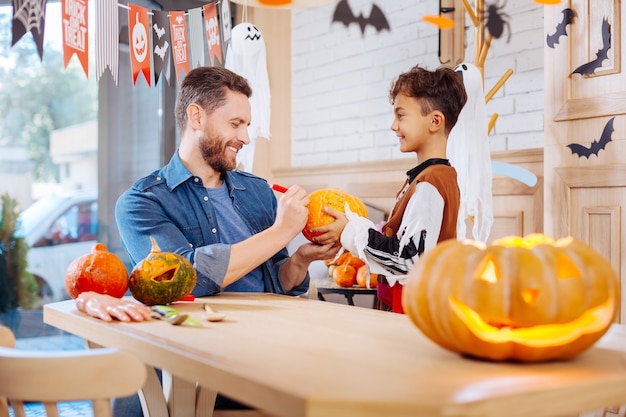 Father helping. Caring bearded father feeling memorable while helping his son wearing Halloween costume painting pumpkin