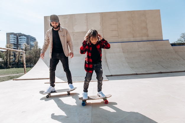 Father and his little son dressed in the casual clothes ride skateboards in a skate park with slides outside at the sunny day .