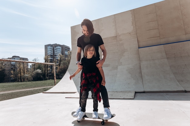 Father and his little son dressed in the casual clothes stand together on the one skateboard in a skate park at the sunny day .