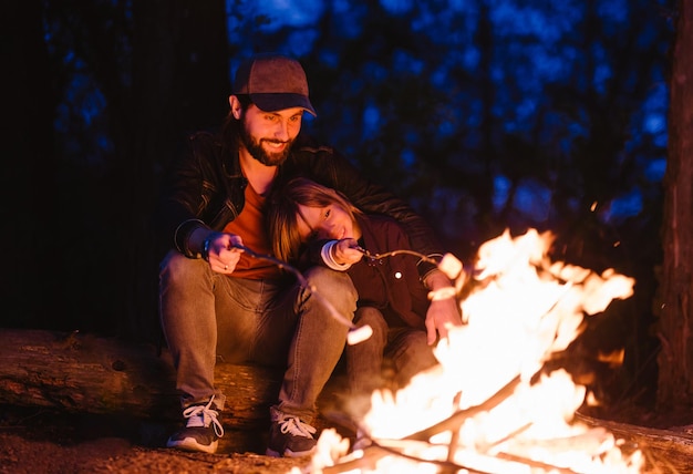 The father and his son sitting on the logs in the forest  and roasting marshmallows on the fire