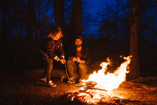 The father and his son sitting on the logs in the forest  and roasting marshmallows on the fire