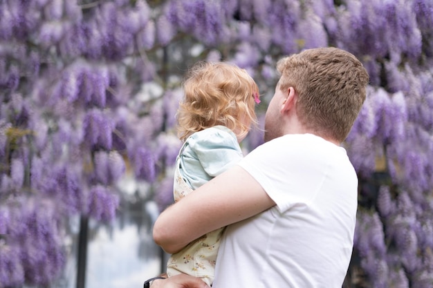 Father holding baby girldaughter outdoor looking at glucinum flowerspurplevery peri wistaria treeyou