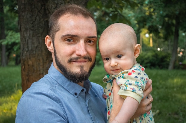 Father holding baby son on the background of a summer landscape close-up. Bearded young man smiling, holding pensive baby boy and looking at camera