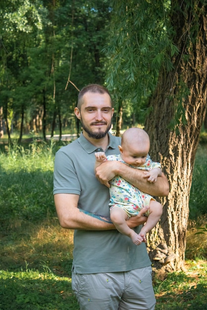 Father holding his baby son on the background of a summer landscape. Bearded young man smiling, holding pensive baby boy and looking at camera