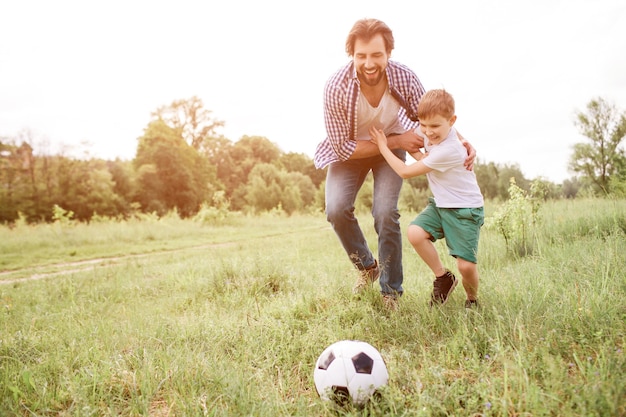 Father is playing football with his son. They are running down the meadow. Boy is hugging his dad and looking at ball. Man is doing the same thing