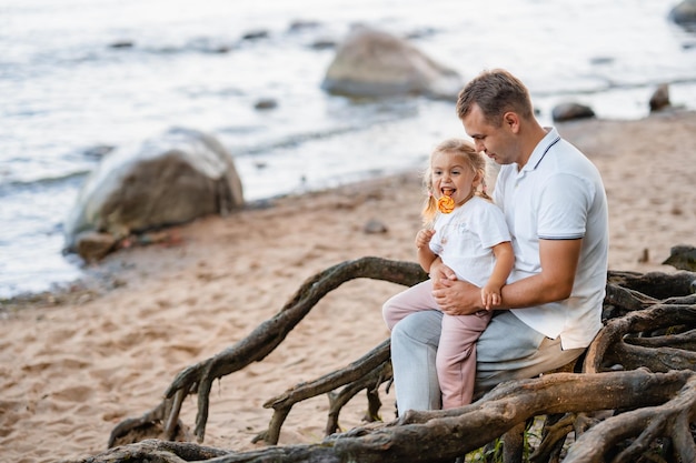 Father and little cute blond daughter together in the summer on the beach in nature a little girl eats a lollipop tree roots