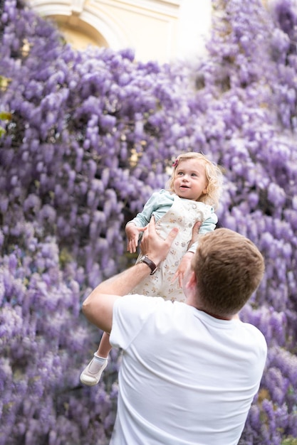 Father playhaving fun outdoor with baby girldaughter on background of glucinum flowerpurplevery peri