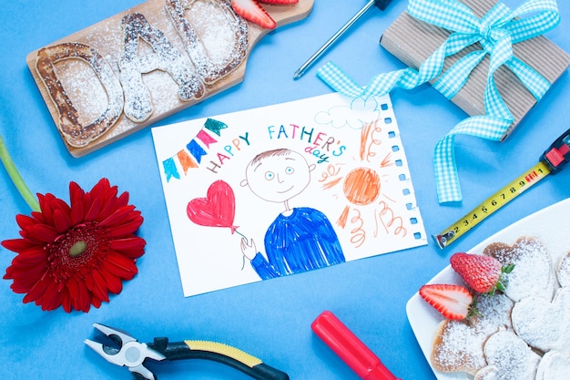 Father's day kid drawing with cake, gift, flowers and more on blue background top view