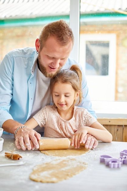 Father showing his little daughter how to roll dough for cookies, baking in kitchen, family cooking at home.