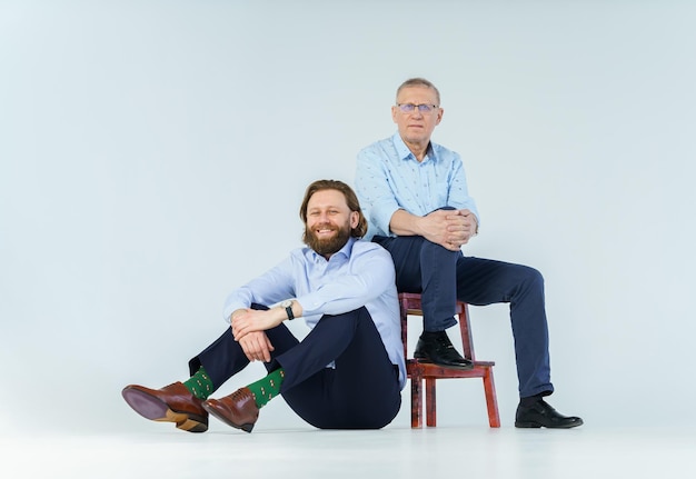 Father sits on the chair and son sits on white background both men look into the camera an elderly