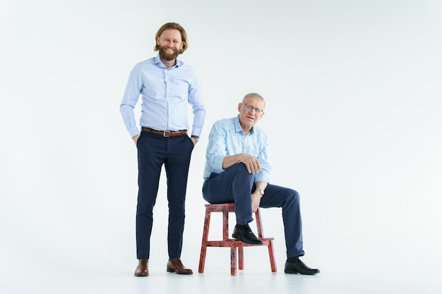 Father sits on the chair and son stands on white background both men look into the camera an elderly