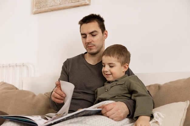 Father and son reading a book on the bed at home. Young attractive man and little boy resting in bedroom. Natural earth colors. Cozy environment. Father reads a fairy tale for his child.