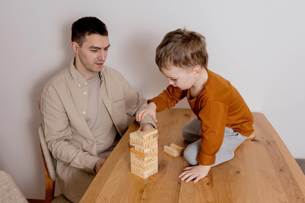 Father and son sitting together at home and playing with wooden blocks Jenga game Little boy and his father spending time together Family time Leisure activity indoors