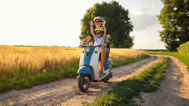 The father and son spending time together riding through the field by pathway on the retro scooter in the sunset