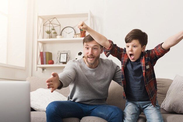 Father and son watching football on TV at home. Emotional man and little boy cheering their favorite team, family enthusiasm, copy space