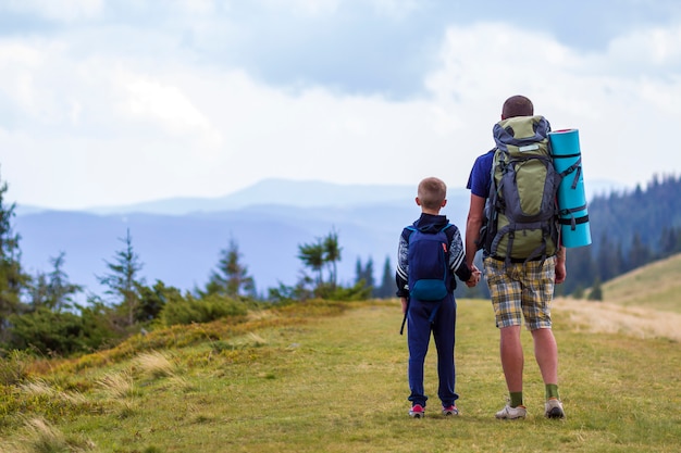 Father and son with backpacks hiking together in scenic summer green mountains. Dad and child standing enjoying landscape mountain view. Active lifestyle, family relations, weekend activity concept.