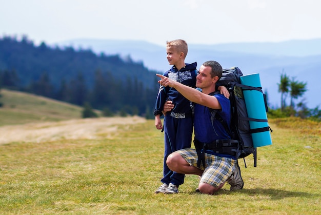 Father and son with backpacks hiking together in scenic summer green mountains. Dad and child standing enjoying landscape mountain view. Active lifestyle, family relations, weekend activity .