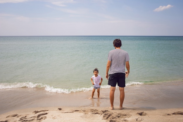 Father in a striped T-shirt walks with his son in shorts and a T-shirt walking on beach in summer during a vacation