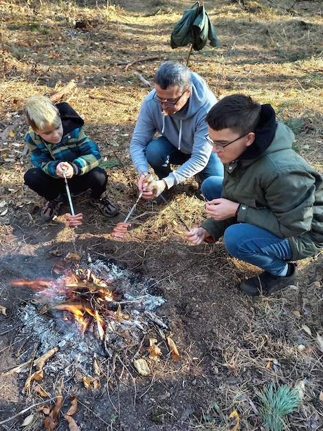 A father and two sons are sitting by the fire and roasting sausages on skewers