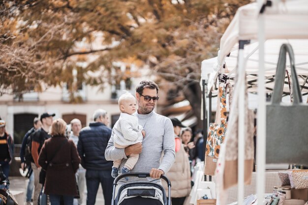 Father walking carrying his infant baby boy child and pushing stroller in crowd of people wisiting sunday flea market in Malaga Spain