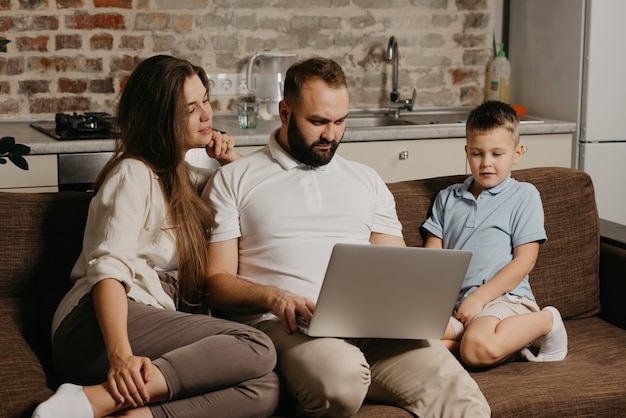 A father with a beard is working remotely on a laptop while his son and wife are staring at the screen