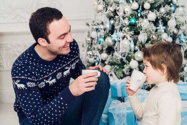 Father with his son sitting near Christmas tree and drinking hot tea