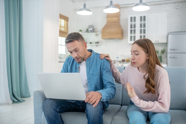 A father with a laptop and a daughter who put her hand on her fathers shoulder, both sitting on the couch.