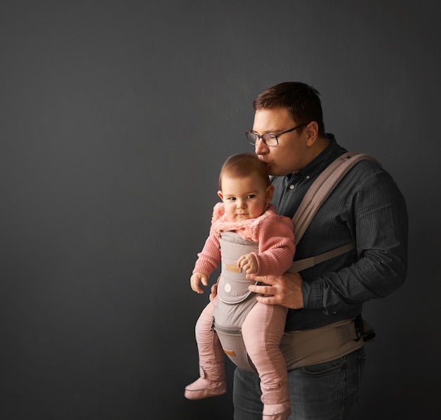 Fathre and his kid in baby carrier at the grey background wall, baby wearing in parenthood