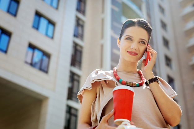 Favourite drink. Delighted joyful woman smiling while holding a cup of coffee