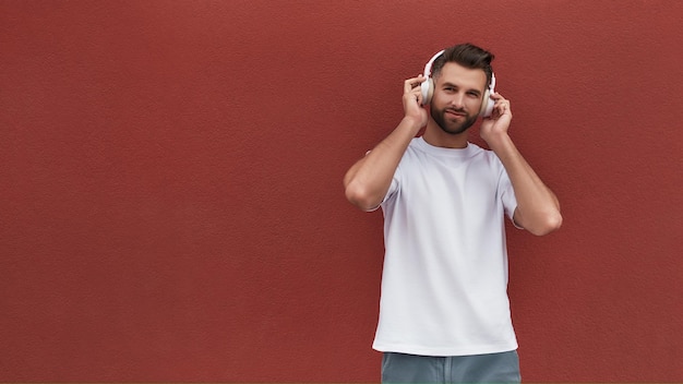 Photo favourite playlist portrait of handsome man in headphones listening to the music and looking at camera while standing against red wall outdoors