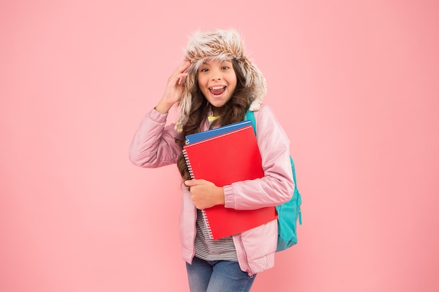 Feel the spirit winter holiday and vacation kid warm clothes pink background pupil daily life schoolgirl backpack and books go home no more exams small happy girl earflap hat back to school