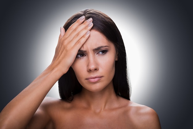 Feeling awful headache. Portrait of frustrated young shirtless woman looking away and touching forehead with hand while standing against grey background