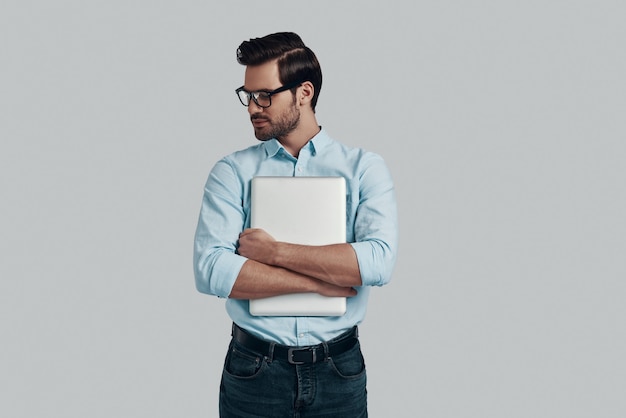 Feeling confident. Handsome young man carrying laptop and smiling while standing against grey background