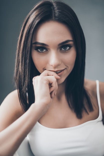 Feeling playful tonight. Attractive young woman holding hand on chin and looking flirty while sitting against grey wall