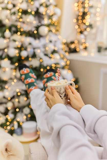 Feet in Santa's socks near the Christmas tree. Woman sitting at the blanket, drinks hot beverage and relaxes warming up their feet in woollen socks. Winter and Christmas holidays concept