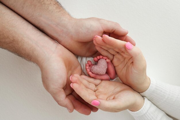 Feet toes heels of a newborn Children39s foot in the hands of mother father parents with a knitted pink heart The legs of the child in the arms of mom and dad On a white background